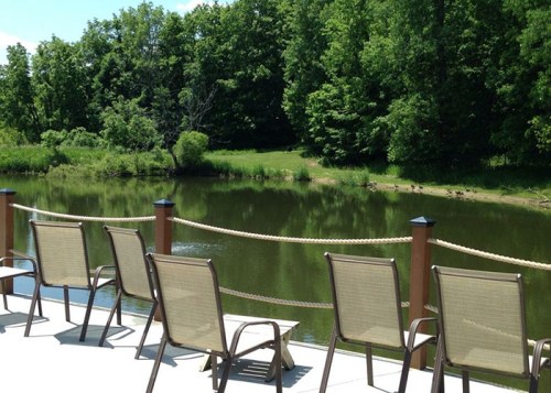 A serene view of a pond surrounded by greenery, with empty chairs lined up along a wooden deck.
