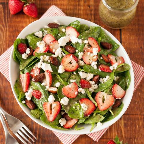 A fresh spinach salad topped with strawberries, almonds, and feta cheese, served in a white bowl on a wooden table.