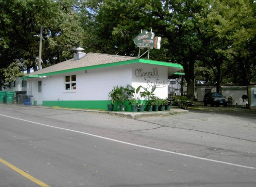 A small, white building with green trim and potted plants outside, set along a tree-lined street.