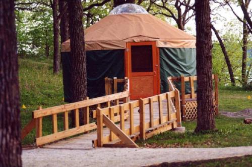 A wooden walkway leads to a round, tan and green yurt nestled among trees in a grassy area.