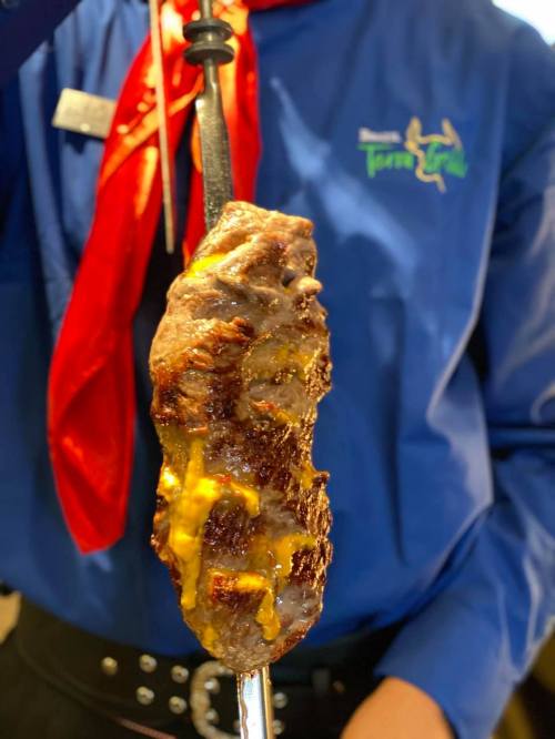 A skewer of grilled meat topped with melted cheese, held by a person in a blue shirt and red scarf.