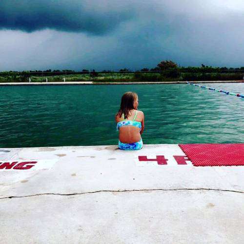 A girl in a swimsuit sits on the edge of a pool, facing dark storm clouds over the water.