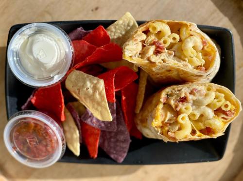 A black container with two burritos filled with macaroni and cheese, surrounded by colorful tortilla chips and dipping sauces.