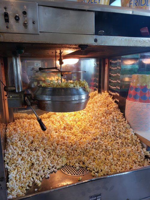 A popcorn machine filled with freshly popped popcorn, with a scoop and colorful cups in the background.