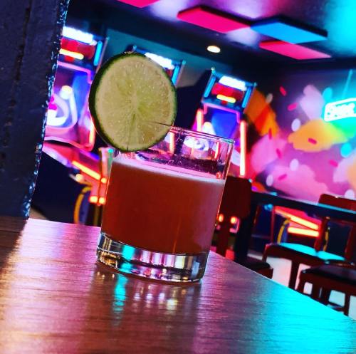 A vibrant cocktail with a lime slice sits on a wooden table, with colorful arcade games in the background.