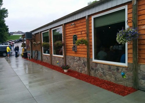 Exterior view of a rustic restaurant with wooden siding, large windows, and flower planters, on a rainy day.