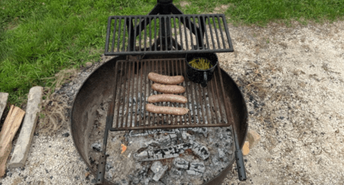 Grill with sausages cooking over hot coals, alongside a pot of green beans. Grass and gravel in the background.