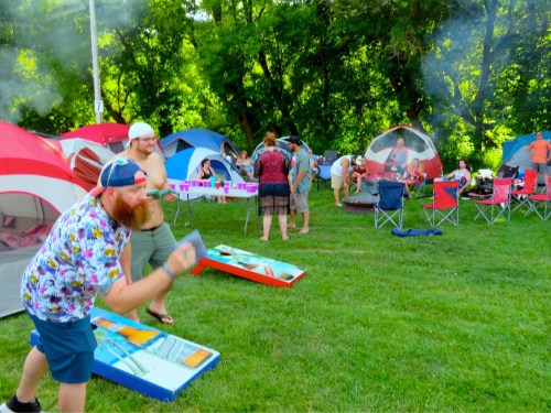 A lively outdoor scene with people playing cornhole, surrounded by colorful tents and green trees.