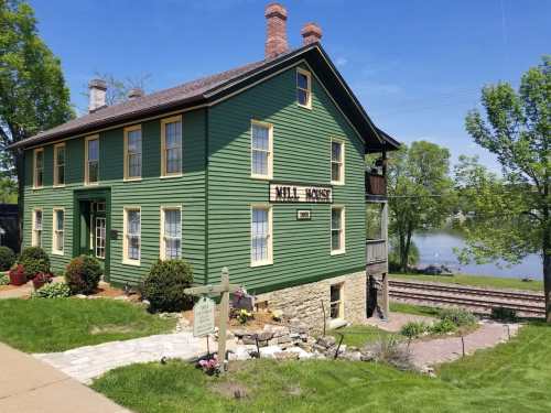 Historic green building with yellow trim, labeled "Mill House," near a river and railway tracks on a sunny day.