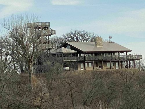 A large, multi-level house on a hill, featuring balconies and a nearby observation tower surrounded by trees.