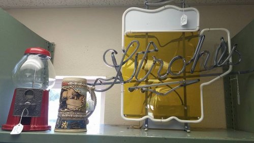 A vintage gumball machine, a decorative beer stein, and a neon Strohs sign displayed on a shelf.