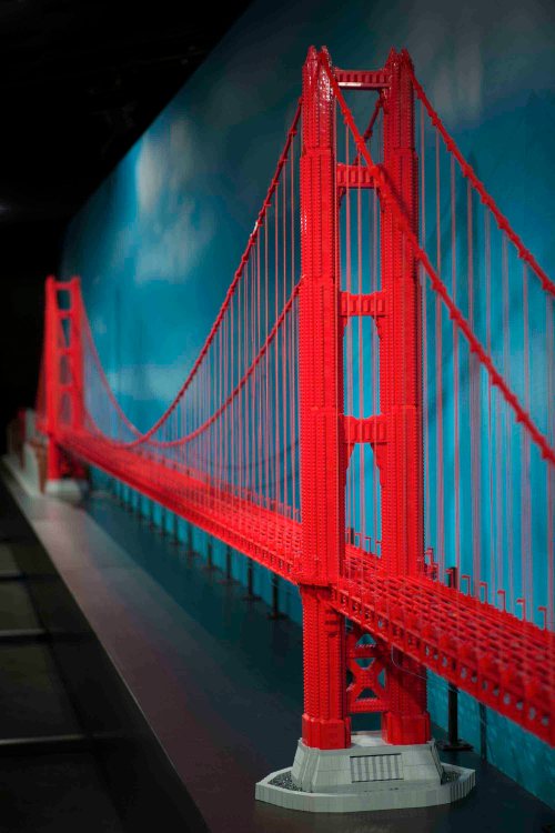 A detailed model of a red suspension bridge made from LEGO bricks, set against a blue sky backdrop.