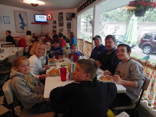 A group of seven people enjoying a meal at a diner, with food and drinks on the table, and a cozy atmosphere.