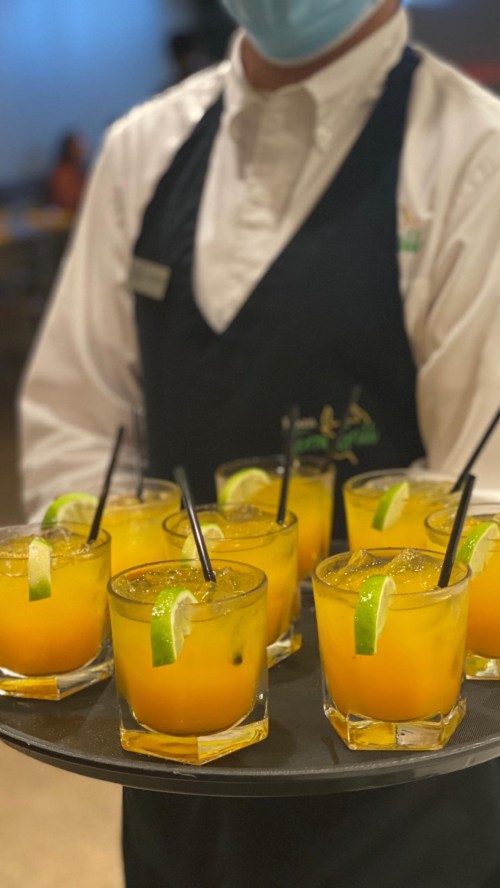 A server in a mask holds a tray of ten orange cocktails garnished with lime slices and straws.