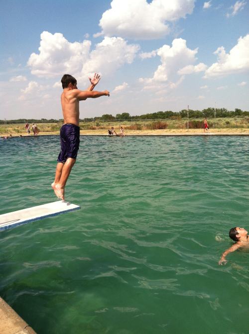 A shirtless boy jumps off a diving board into a green lake, with another person swimming nearby under a cloudy sky.