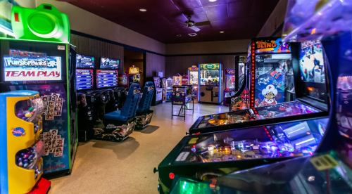 A vibrant arcade filled with various gaming machines, including pinball and racing games, under dim lighting.