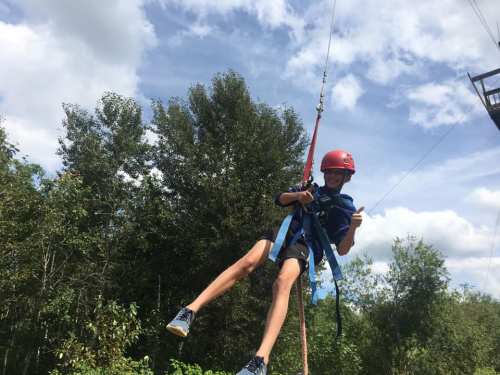 A child in a helmet and harness smiles and gives a thumbs up while zip-lining through a forested area.