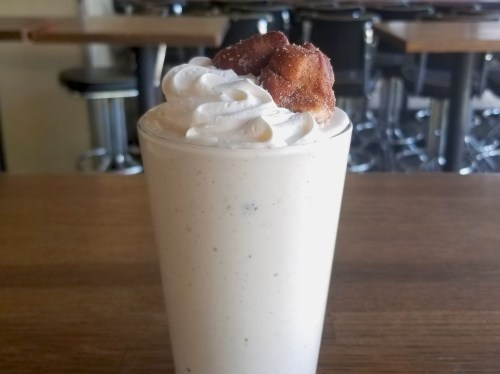 A tall glass of creamy milkshake topped with whipped cream and a piece of fried dough.