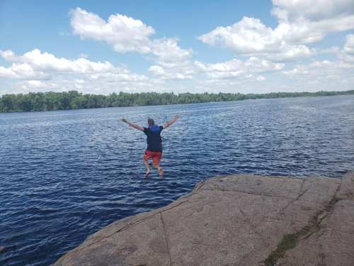 A person jumps off a rocky ledge into a calm lake under a blue sky with fluffy clouds.