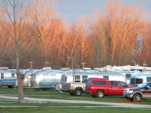 A row of silver Airstream trailers parked in a grassy area, with trees reflecting warm sunset colors in the background.