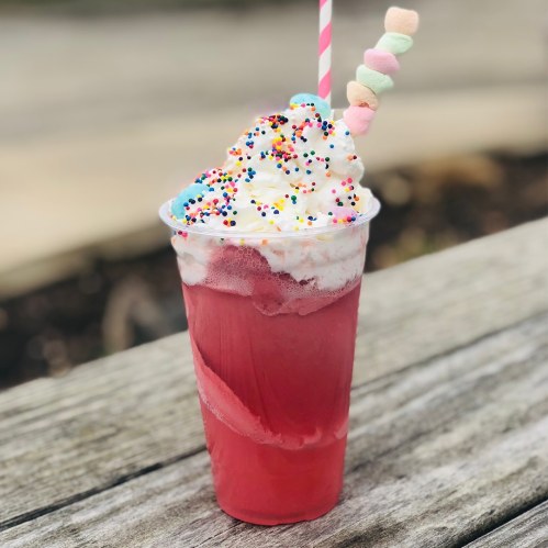 A colorful drink topped with whipped cream, sprinkles, and a marshmallow skewer, placed on a wooden surface.