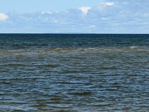 A distant bridge spans across the water, with waves gently rippling in the foreground under a partly cloudy sky.