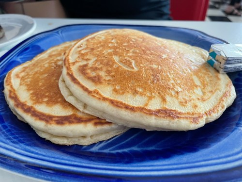 Three golden-brown pancakes stacked on a blue plate, with a small carton of syrup beside them.