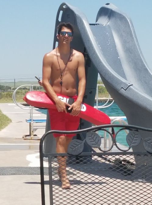 A lifeguard in red shorts and sunglasses stands by a pool, holding a rescue buoy near a water slide.