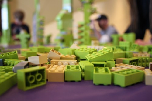 A close-up of scattered green and beige LEGO bricks on a table, with blurred figures in the background.