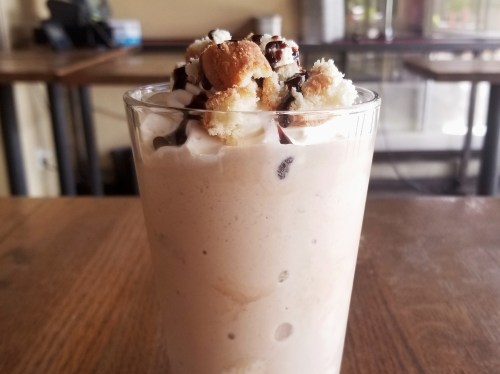A tall glass filled with a creamy dessert topped with whipped cream, chocolate drizzle, and cake pieces.