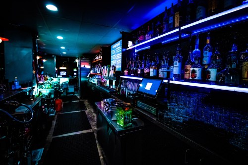 A dimly lit bar with colorful LED lights, shelves lined with various bottles, and a sleek counter.