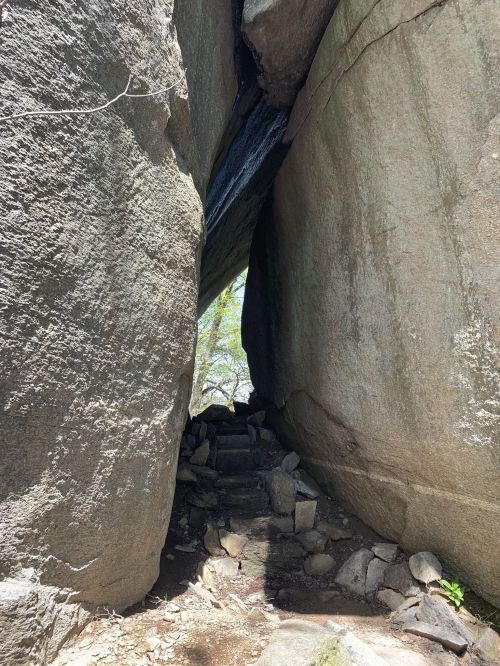Narrow passage between large rocks, leading to a small staircase and greenery in the background.