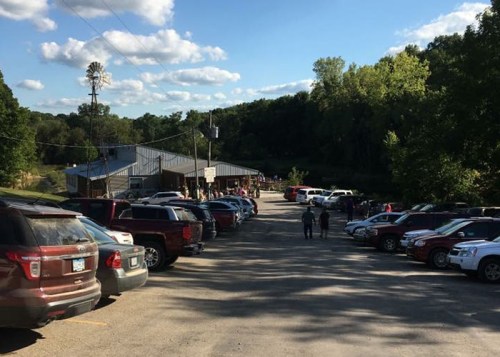 A parking lot filled with cars near a building, surrounded by trees and a clear blue sky with scattered clouds.