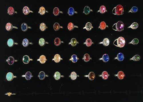 A display of colorful rings featuring various gemstones set in silver, arranged in rows on a black background.