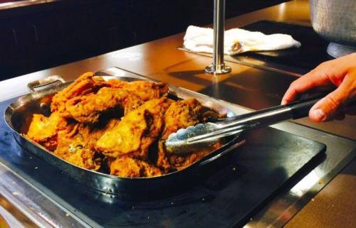 A hand using tongs to serve crispy fried chicken from a metal tray on a countertop.
