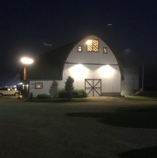 A well-lit barn at night, featuring a large window and surrounded by greenery and parked cars.