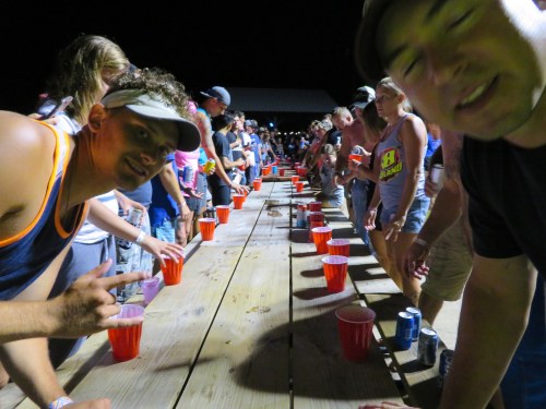 A crowded outdoor party scene with people lined up at a long table, each with red cups, enjoying a lively atmosphere.