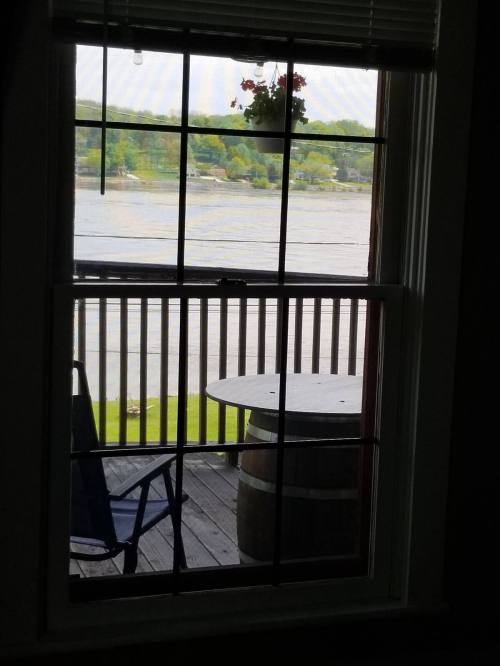 View from a window showing a wooden deck, a chair, and a scenic river landscape with greenery in the background.