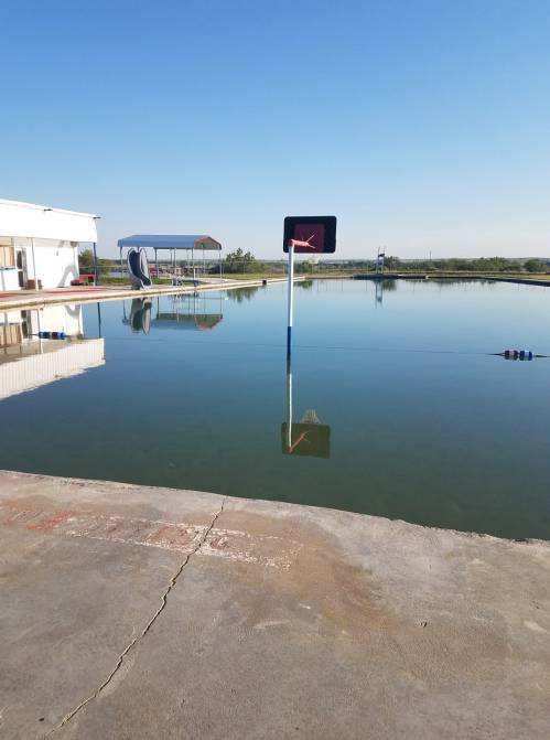 A calm pool reflecting the sky, with a basketball hoop at the edge and a slide in the background.