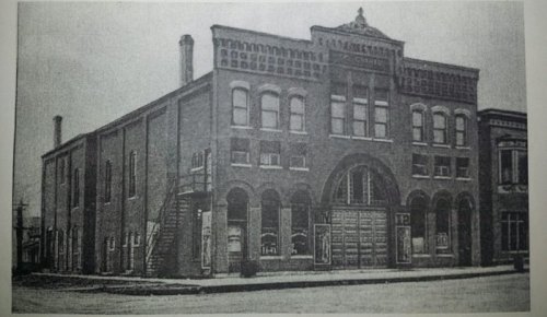 Historic brick building with arched windows and a staircase, set in a street scene from the past.