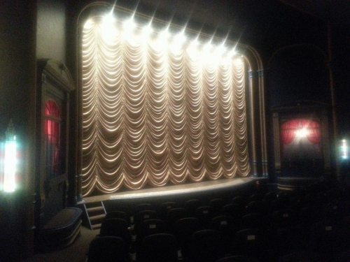 A dimly lit theater stage with a golden curtain and empty seats in the foreground.