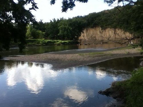 A serene river scene with a sandy bank, lush greenery, and a rocky cliff reflecting in the calm water.