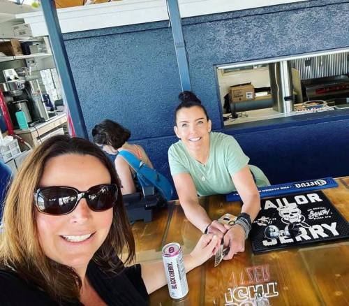 Two women smiling at a table in a casual setting, one holding a drink, with a bar in the background.