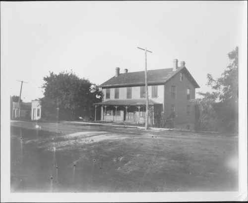 A historic two-story house with a porch, surrounded by trees and utility poles, on a dirt road. Black and white photo.