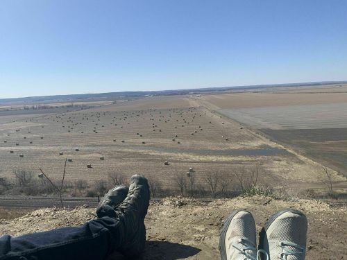 Two pairs of feet resting on a ledge, overlooking a vast, open field with hay bales scattered across the landscape.