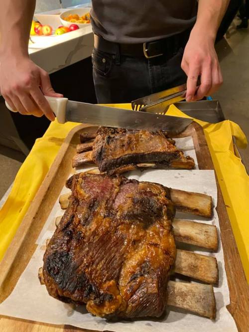 A person slices a large, grilled rib roast on a wooden platter, with fresh apples in the background.