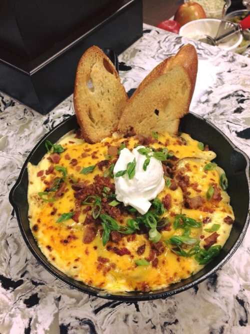 A skillet filled with cheesy baked macaroni topped with bacon, green onions, sour cream, and two slices of toasted bread.