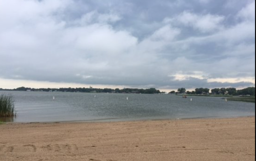 A sandy beach by a calm lake under a cloudy sky, with distant trees and boats visible on the water.