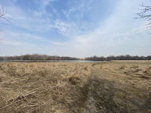 A wide view of a grassy field leading to a calm water body, surrounded by trees under a cloudy sky.