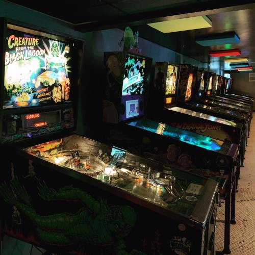 A row of vintage pinball machines, including "Creature from the Black Lagoon" and "Indiana Jones," in a dimly lit arcade.